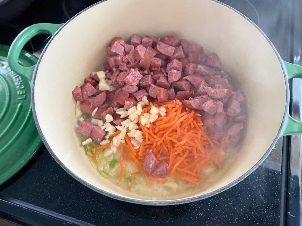 White ceramic pot filled sausage, garlic, carrots, and liquid on a black stove top.