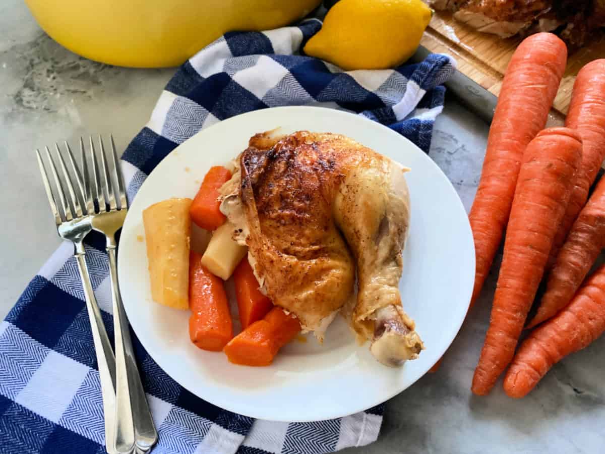 White plate filled with roasted chicken leg and vegetables on a blue and white checkered cloth.