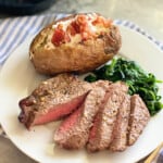 Sliced steak, baked potatoe, and spianch on a white plate sitting on a white and blue striped cloth.