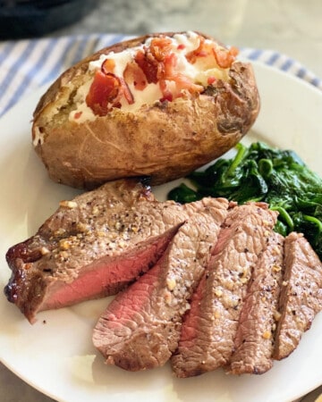 Sliced steak, baked potatoe, and spianch on a white plate sitting on a white and blue striped cloth.