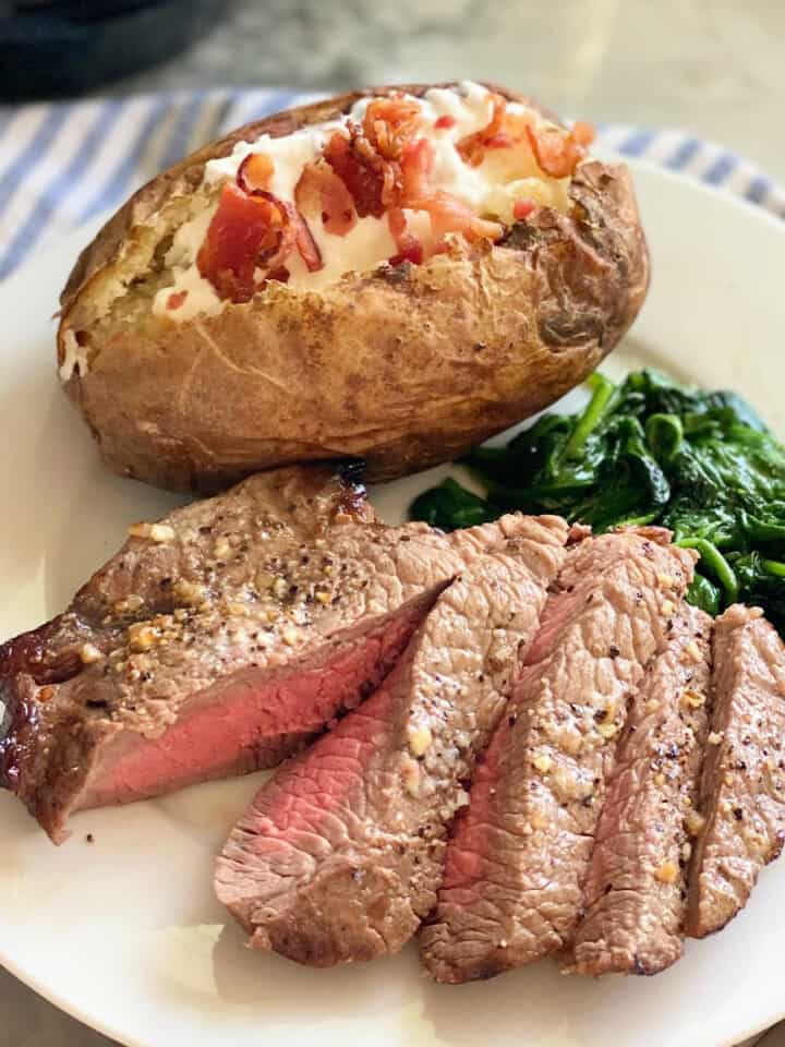 Sliced steak, baked potatoe, and spianch on a white plate sitting on a white and blue striped cloth.