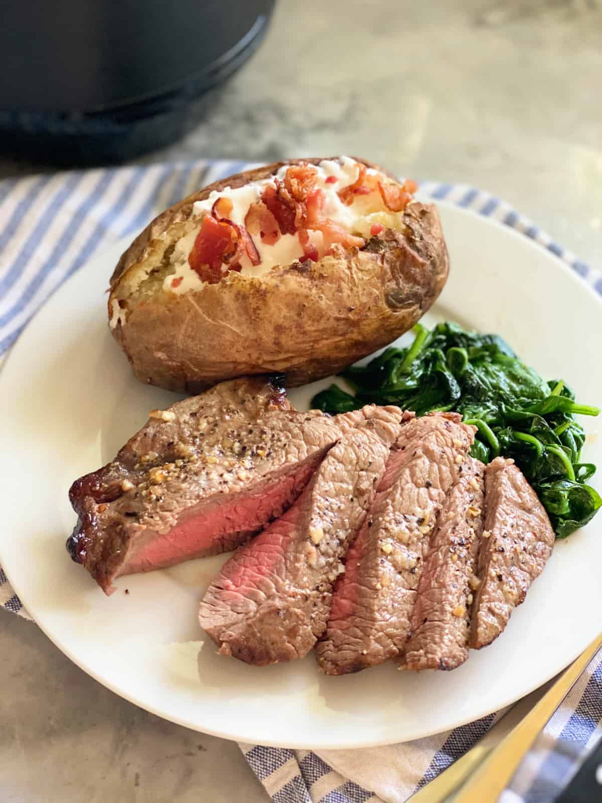 Sliced steak with baked potato and spinach on a white plate and blue and white cloth next to it.