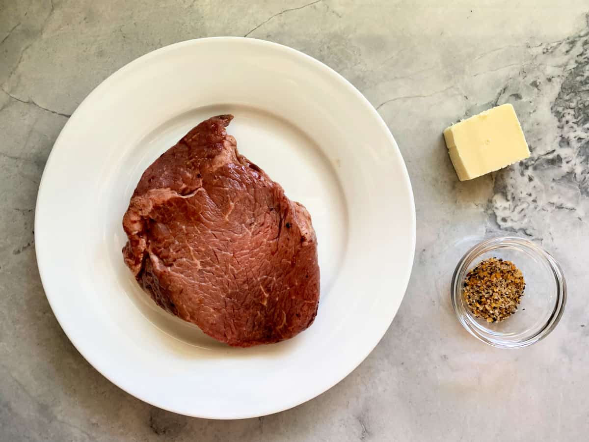 Steak on white plate, square of butter, and seassoning in dish on a grey marble countertop.
