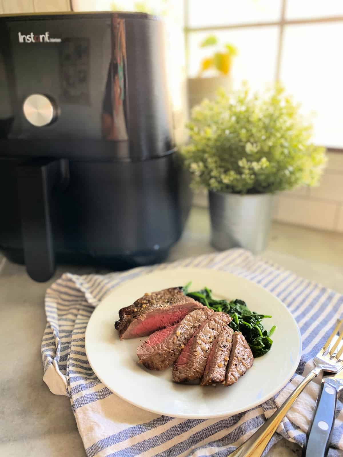 Pieces of sliced steak with sauted spinach on a white plate with an Air Fryer in the background.