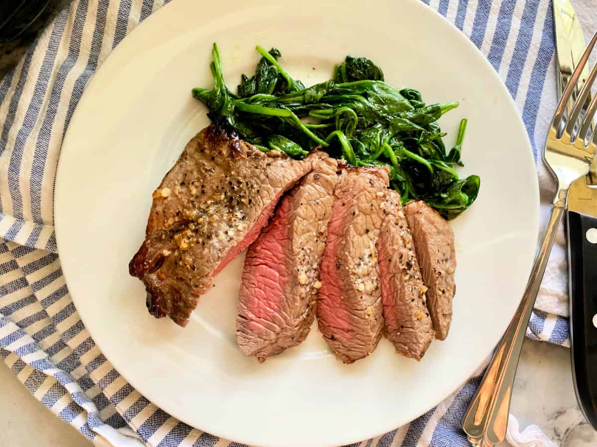 Four pieces of sliced steak with sauteed spinach with a fork and steak knife next to it.