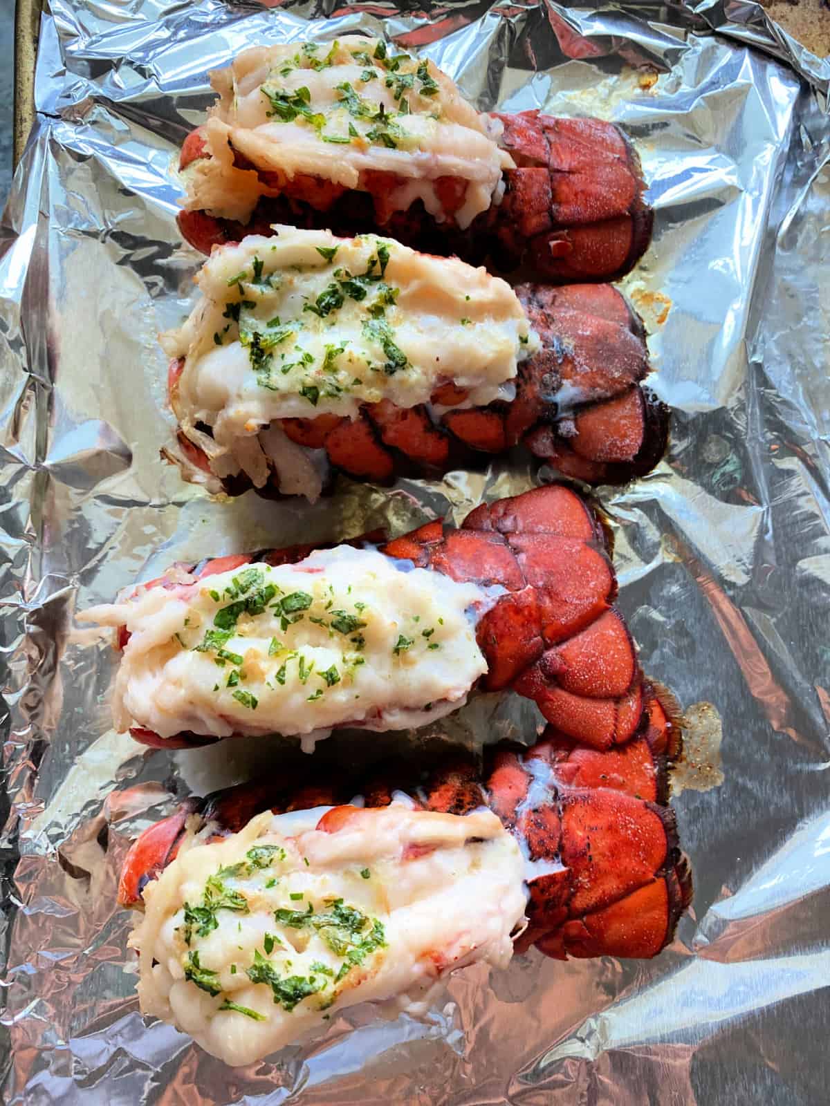 Four cooked lobster tails on an aluminum foil covered tray with herbs on top of the lobster meat.