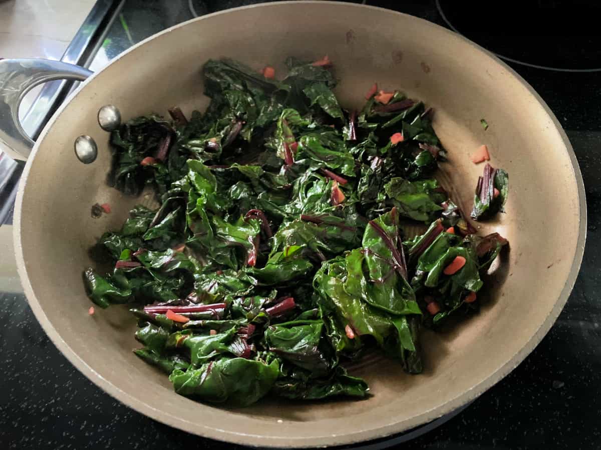 Brown frying pan filled with chopped greens and garlic on a black stove top.