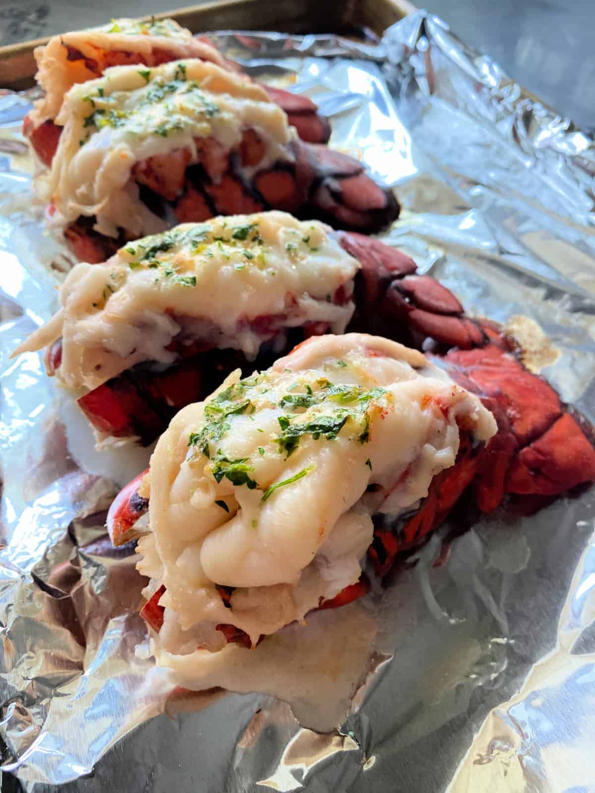 Four lobster tails topped with fresh herbs on a baking sheet.