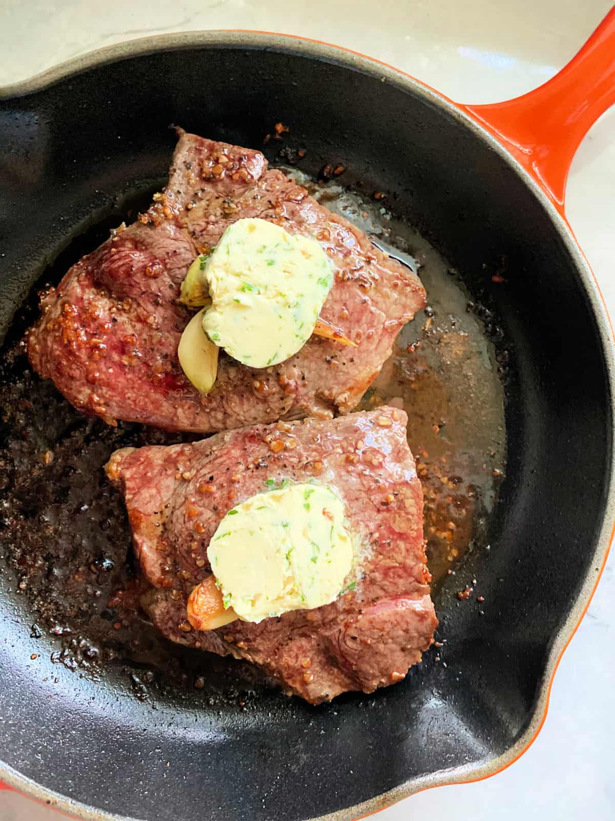 Two steaks in a cast iron skillet with compound butter on top.
