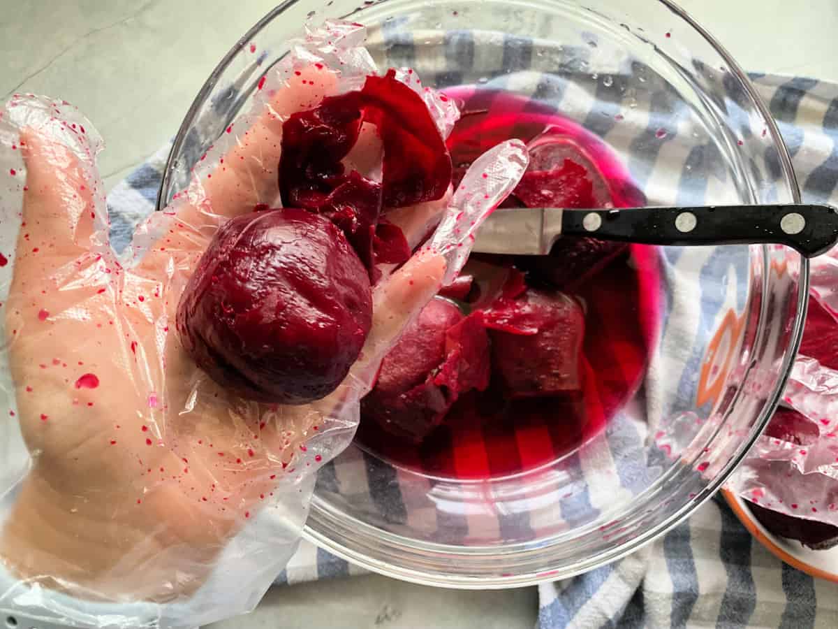 Hand in glove holding a beet with skin peeled back over a bowl of beets in water.