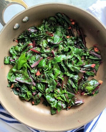 Close up of beet greens in a frying pan on a white and blue striped cloth.