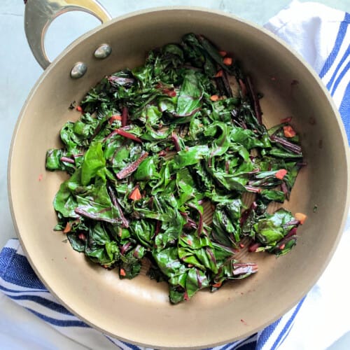 Close up of beet greens in a frying pan on a white and blue striped cloth.
