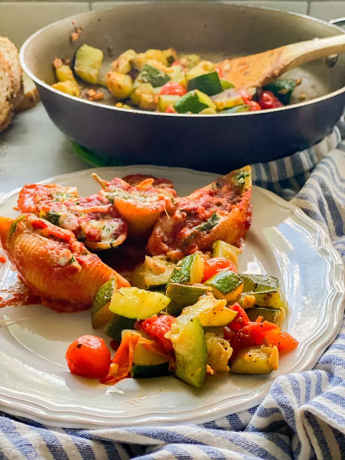 Plate of sautéed zucchini and tomatoes with stuffed shells, with a pan of more diced zucchini and tomatoes in the background.