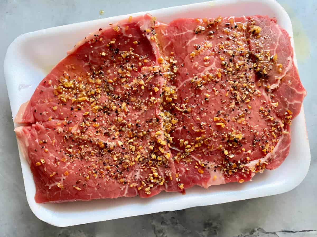 Two steaks in a white tray with seasoning on top.