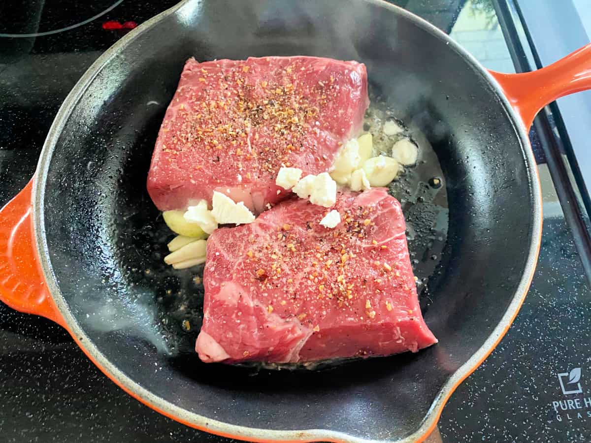 Orange cast iron skillet on stove top with steak, garlic, and butter.