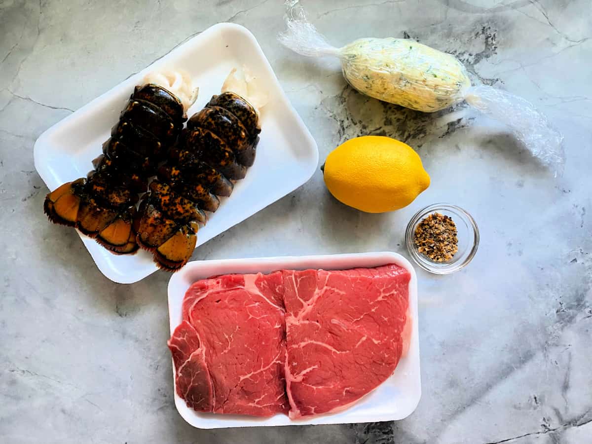 Ingredients on counter: lobster tails, steak, lemon, compound butter, and seasoning in a glass bowl.