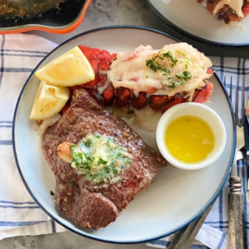 White and blue plate with a steak with butter, lobster, lemon wedges and drawn butter.
