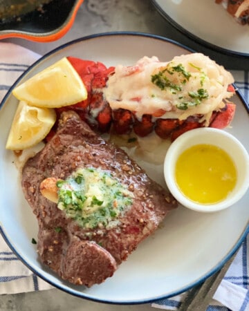 White and blue plate with a steak with butter, lobster, lemon wedges and drawn butter.