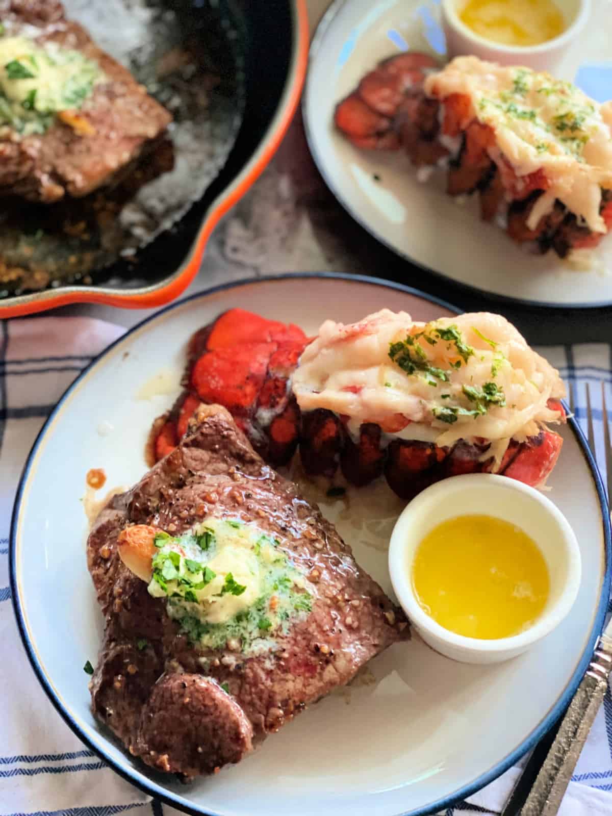 Steak with compound butter, lobster tail, and drawn butter in a little cup with another plate behind it.