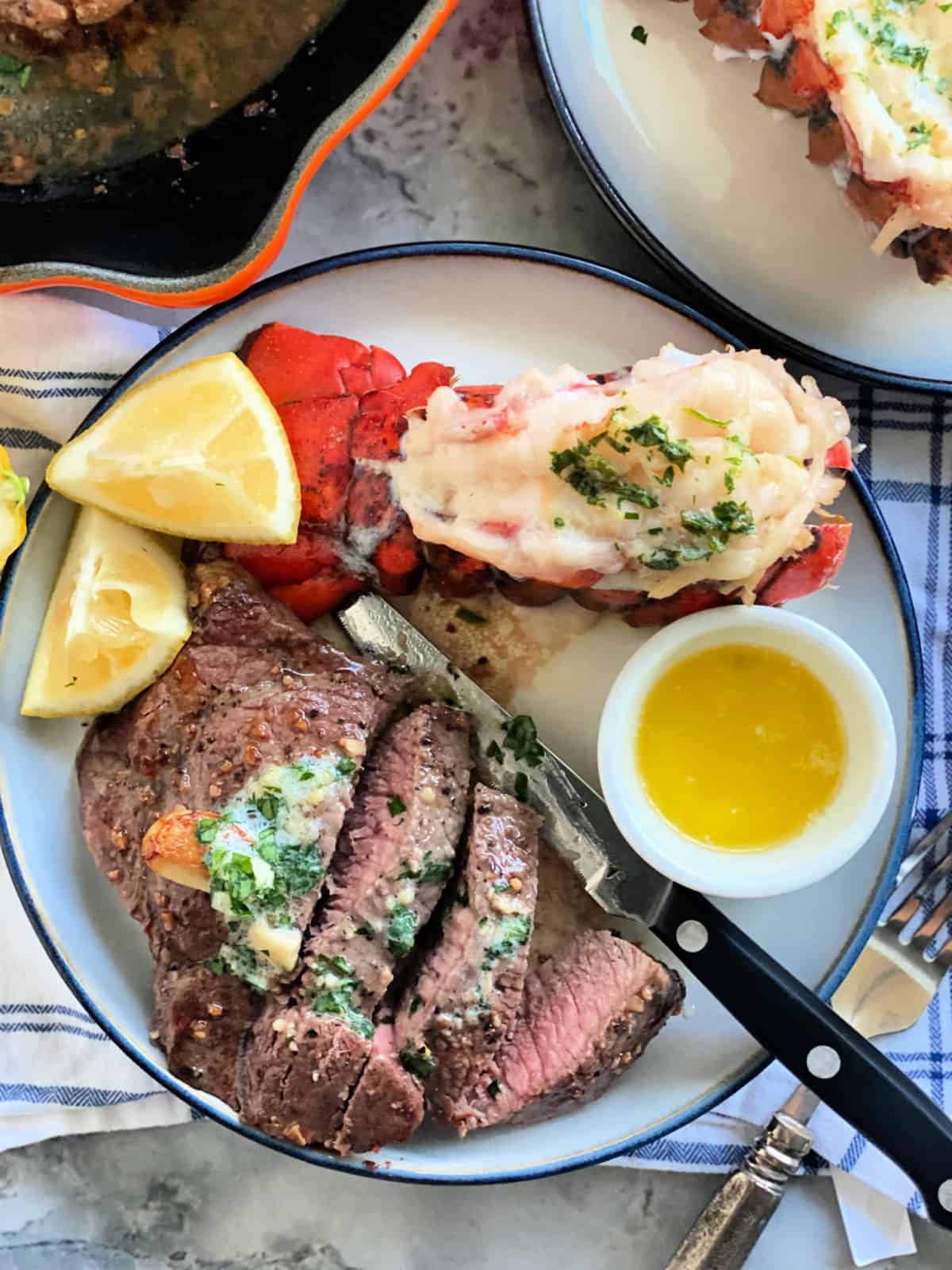 Sliced steak with a steak knife next to a lobster tail with lemon wedges and drawn butter.