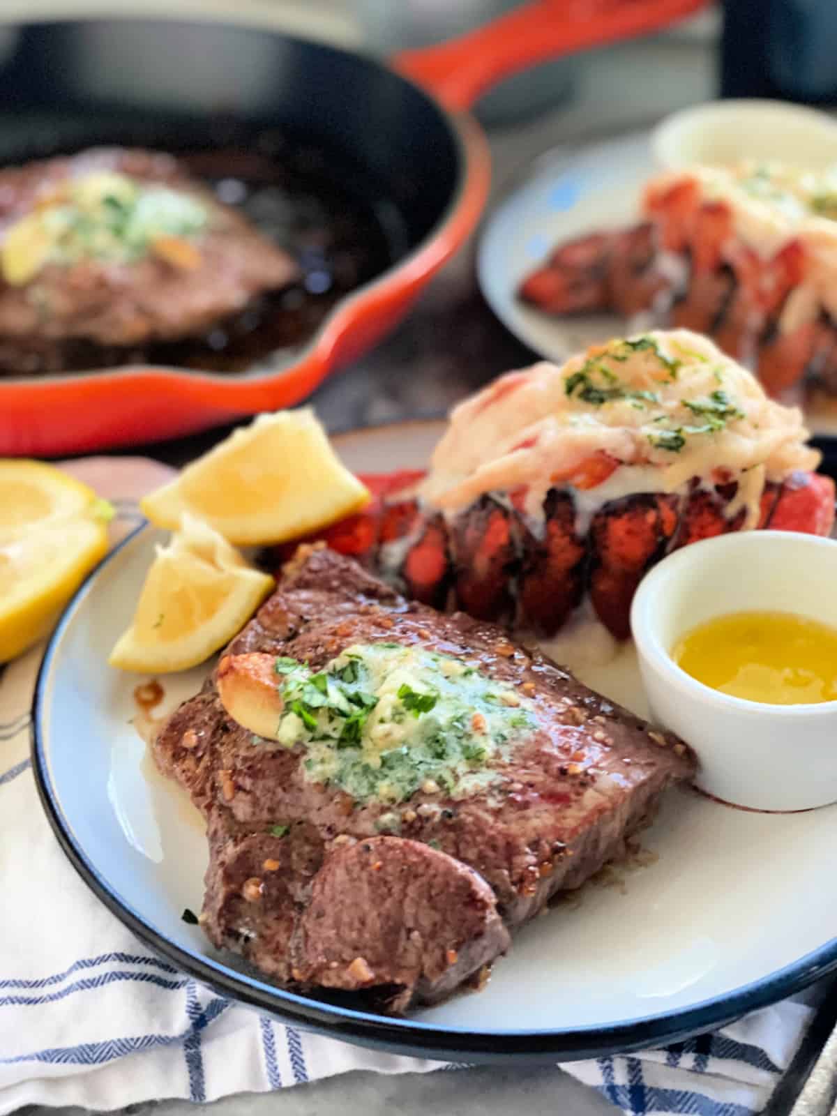 Steak with compound butter and herbs with lemon wedge and lobster tails in background.