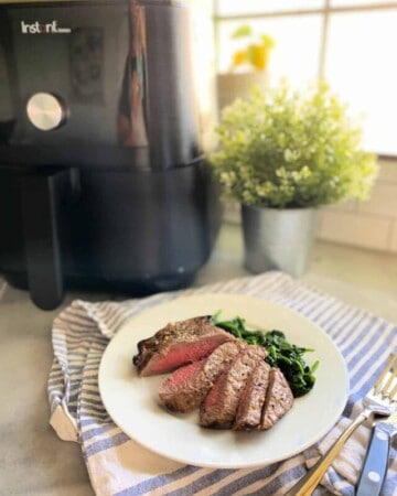 Pieces of sliced steak with sauted spinach on a white plate with an Air Fryer in the background.