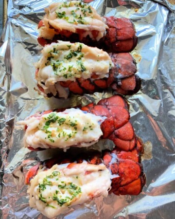 Four cooked lobster tails on an aluminum foil covered tray with herbs on top of the lobster meat.