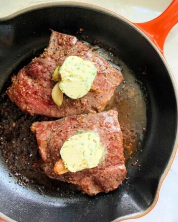 Two steaks in a cast iron skillet with compound butter on top.