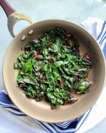 Brown frying pan with sauteed greens in it on a marble countertop.