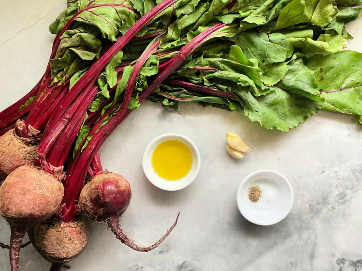 Fresh bundle of beets, olive oil, garlic, salt, and pepper on a marble countertop.