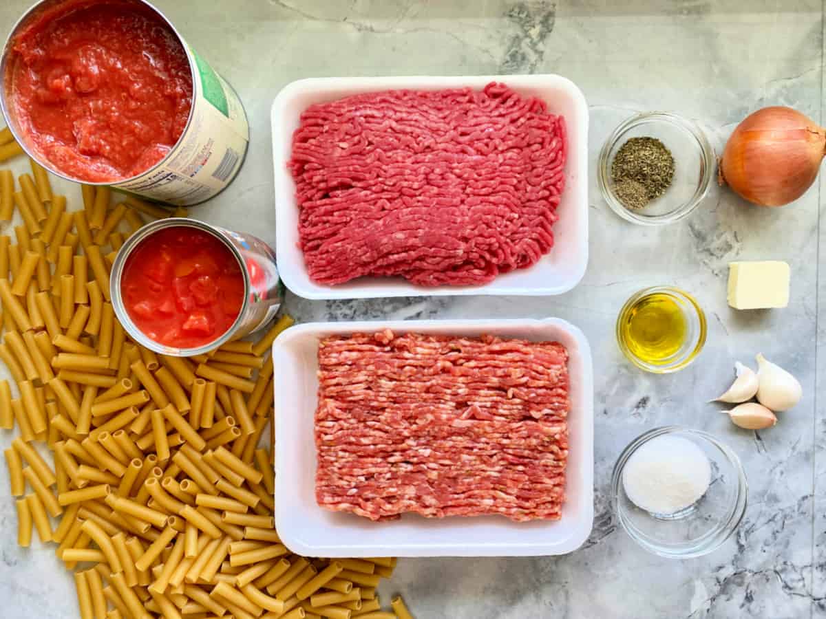 Ingredients on counter; groundbeef, ground pork, penne pasta, canned tomatoes, onion, garlic sugar, olive oil, and spices.