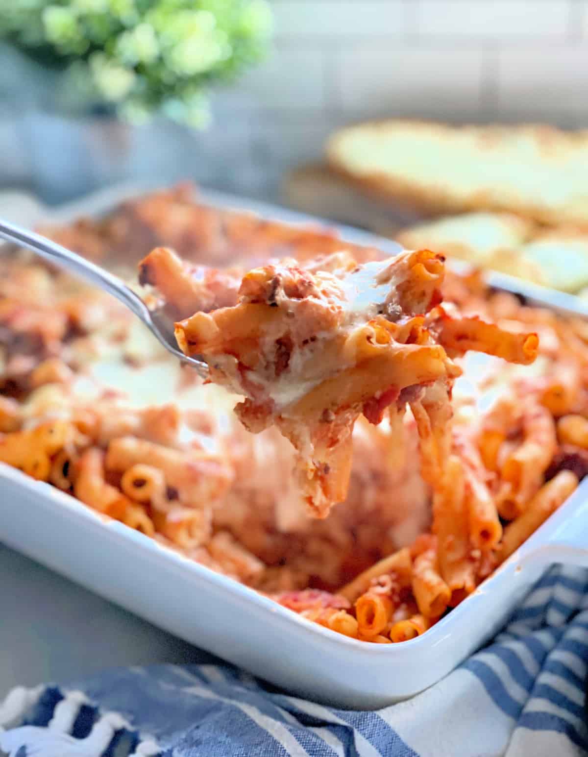 Spoon pulling ziti noodles and cheese out of a white casserole dish.