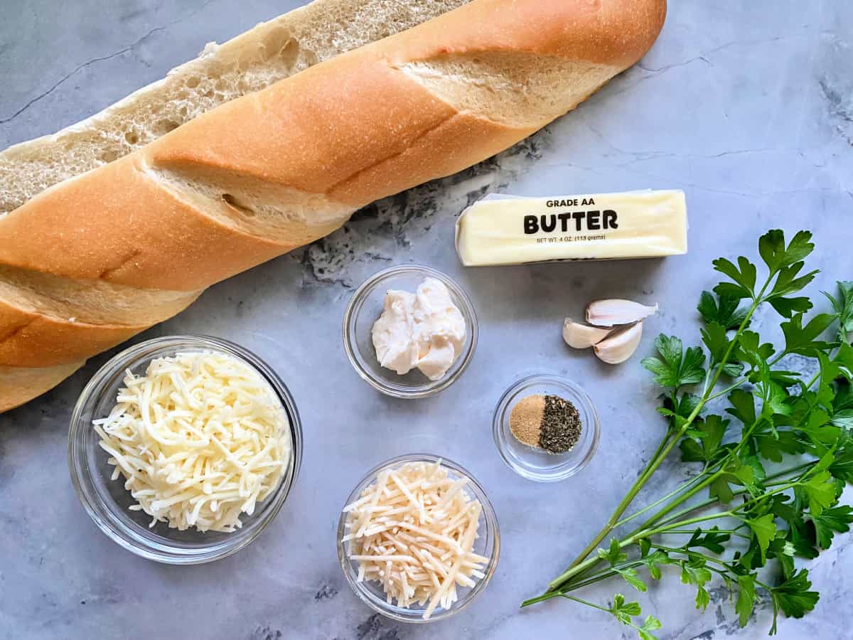 Ingredients on counter: French bread, butter, mayonnasie, garlic, italian seasoning, garlic powder, fresh parsley, mozzarella, and grated parmesan.