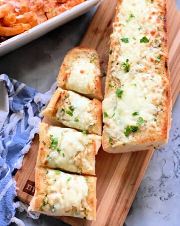 Two loaves of garlic cheese bread with one cut into 4 pieces resting on a wood cutting board.