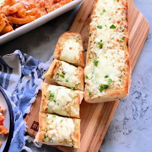 Two loaves of garlic cheese bread with one cut into 4 pieces resting on a wood cutting board.
