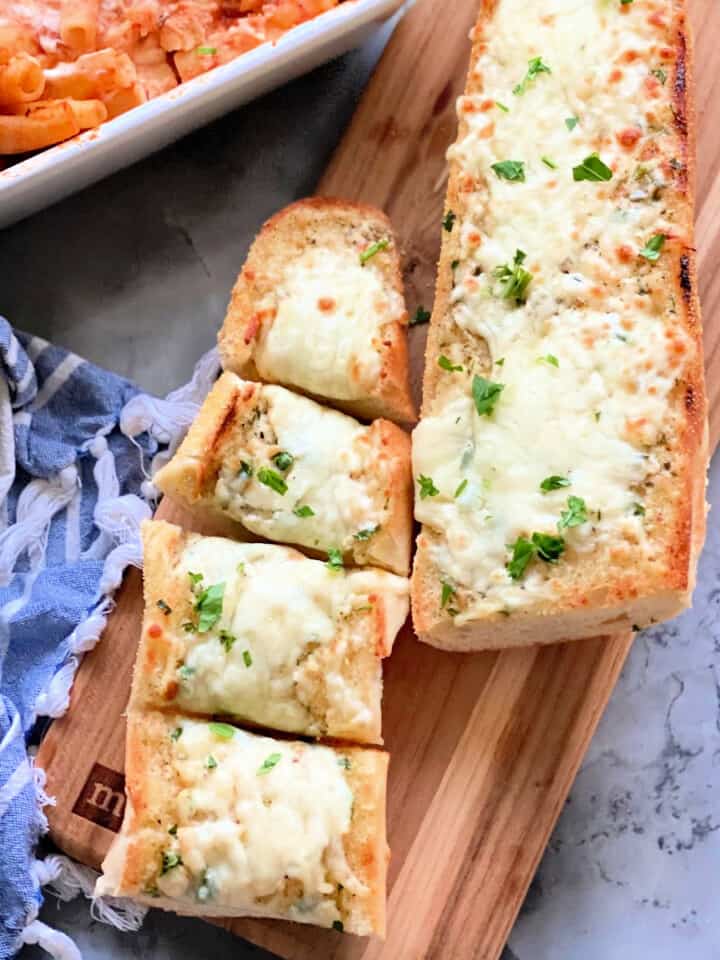 Two loaves of garlic cheese bread with one cut into 4 pieces resting on a wood cutting board.