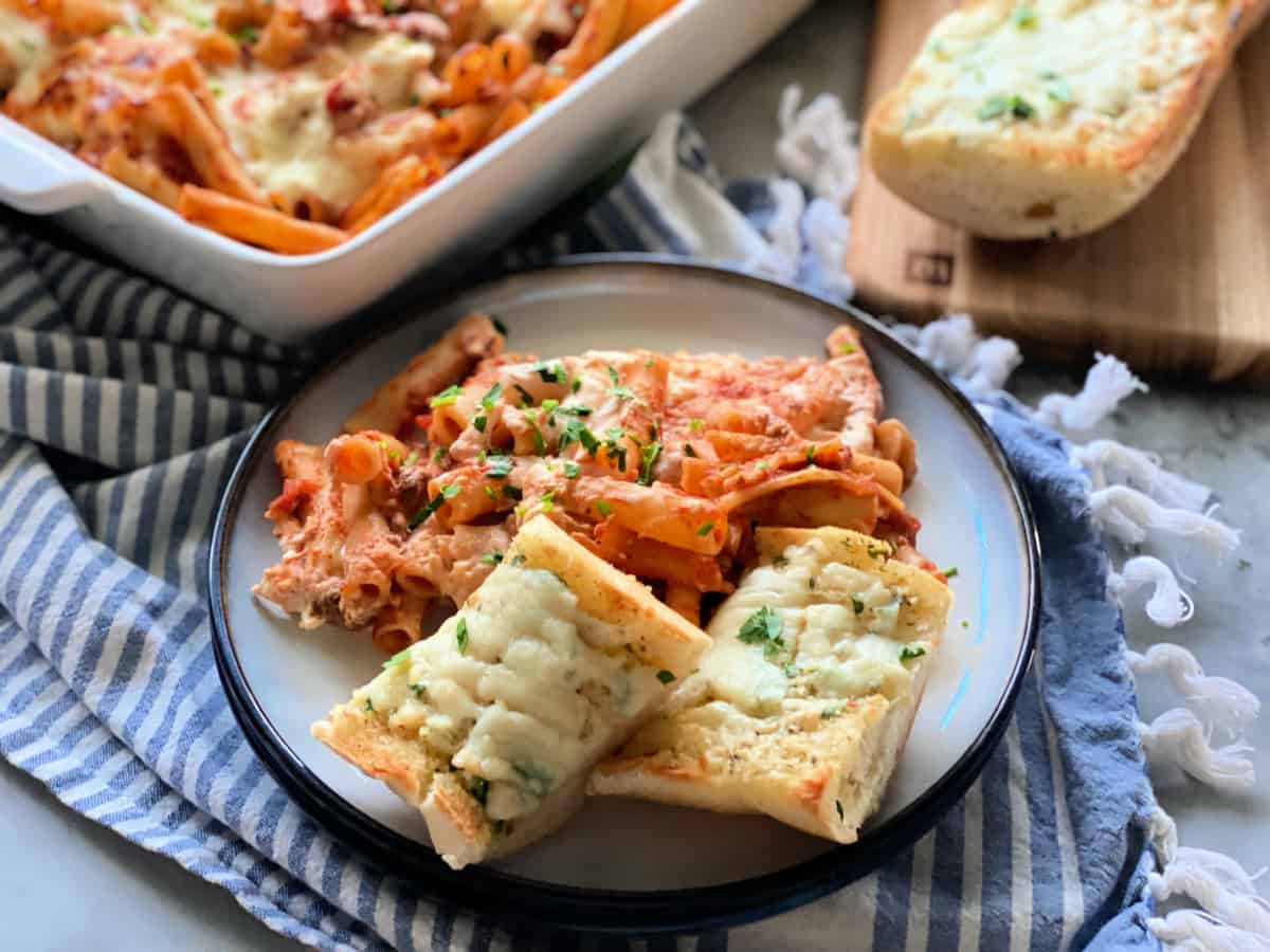 Round plate filled with two slices of cheese bread with baked ziti and baked ziti in the background.