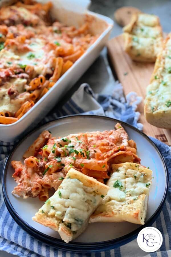 White plate filled with ziti and cheese bread with pan of baked ziti in background with logo on right corner.