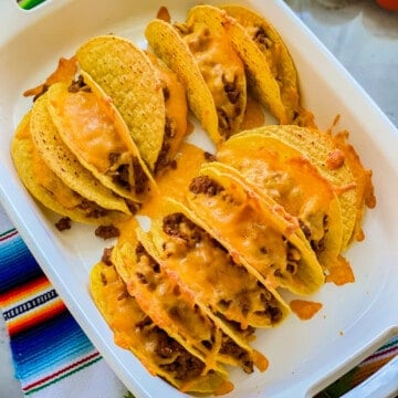 Hard taco shells in a white baking dish filled with meat and cheese.
