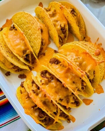 Hard taco shells in a white baking dish filled with meat and cheese.