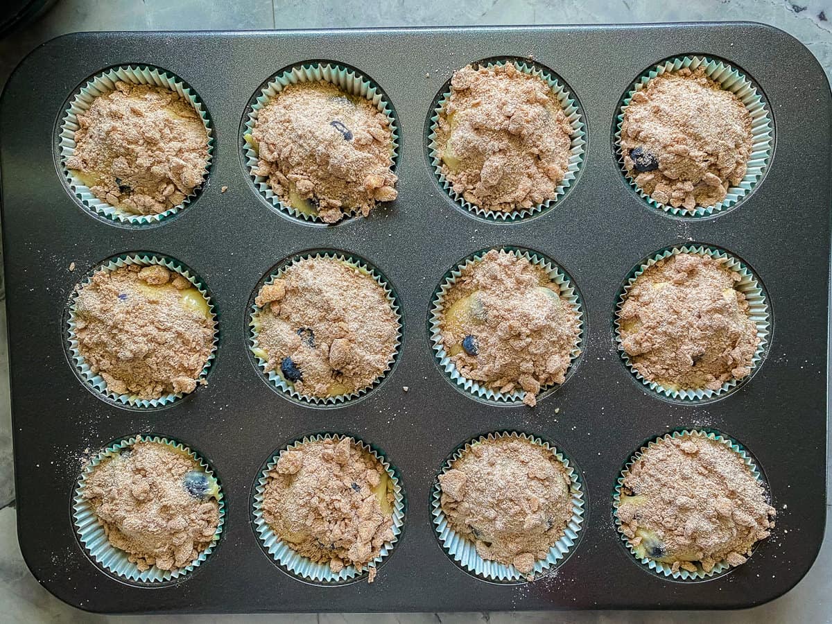Blueberry muffins with crumb toppings on top resting in a muffin pan.