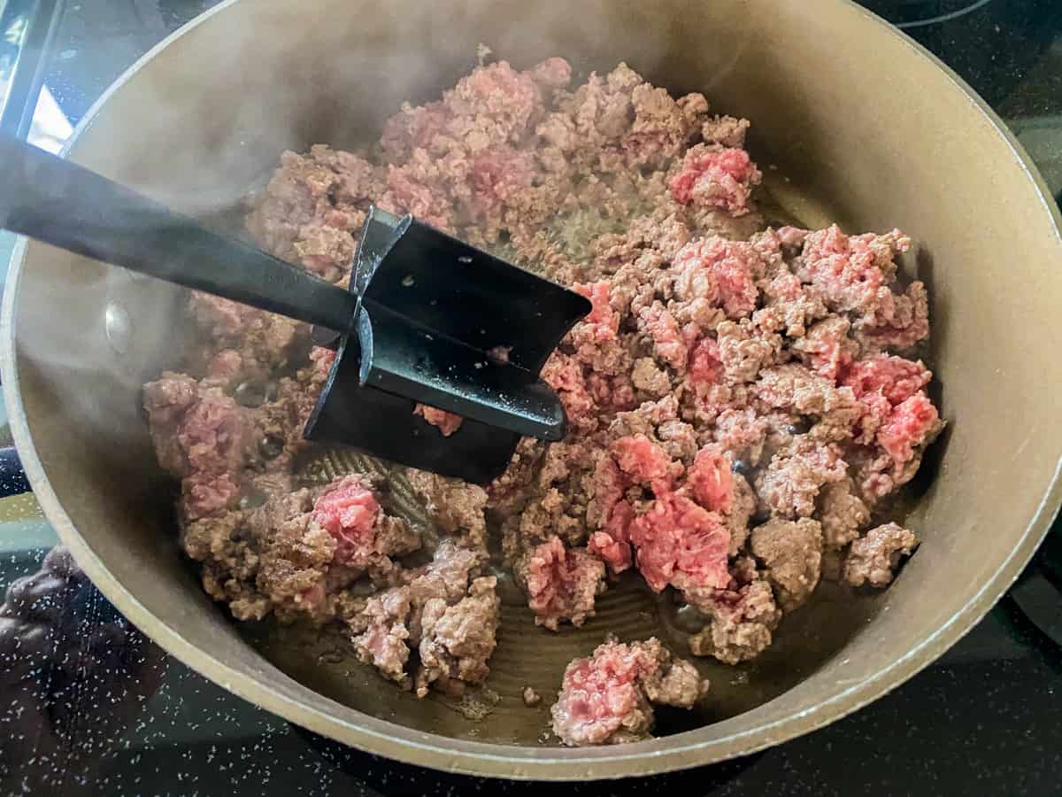 Ground beef being cooked in a pot and being mashed by a masher.