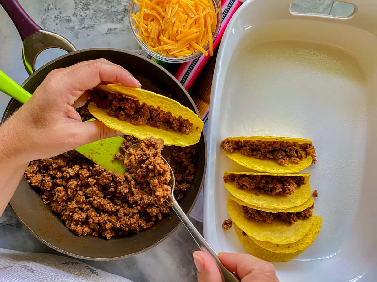 Beef tacos in a baking dish with a taco being filled with the ground beef that was cooked in a pot.