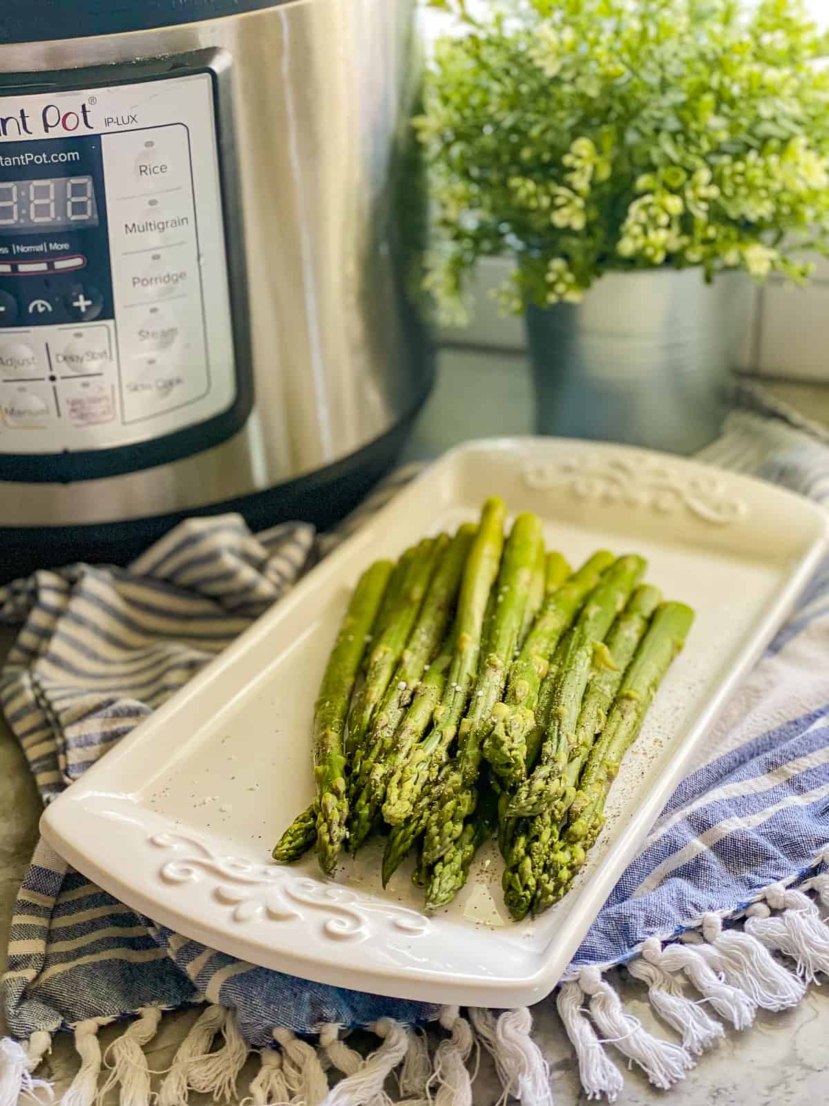 Cooked asparagus resting on a white serving dish that's laying on top of a blue and white striped kitchen towel, with the Instant pot in the background.