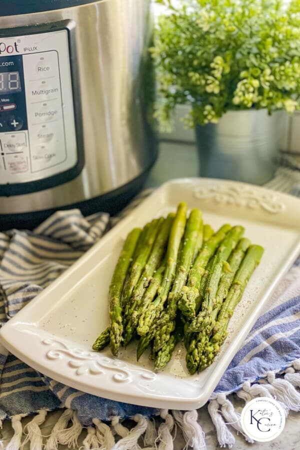 Cooked Asparagus resting on a white serving dish with an instant pot in the background. The Katie's Cucina logo is seen in the bottom right corner.