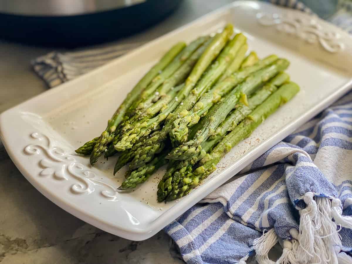 Cooked asparagus resting on a white serving dish that's laying on top of a blue and white striped kitchen towel.