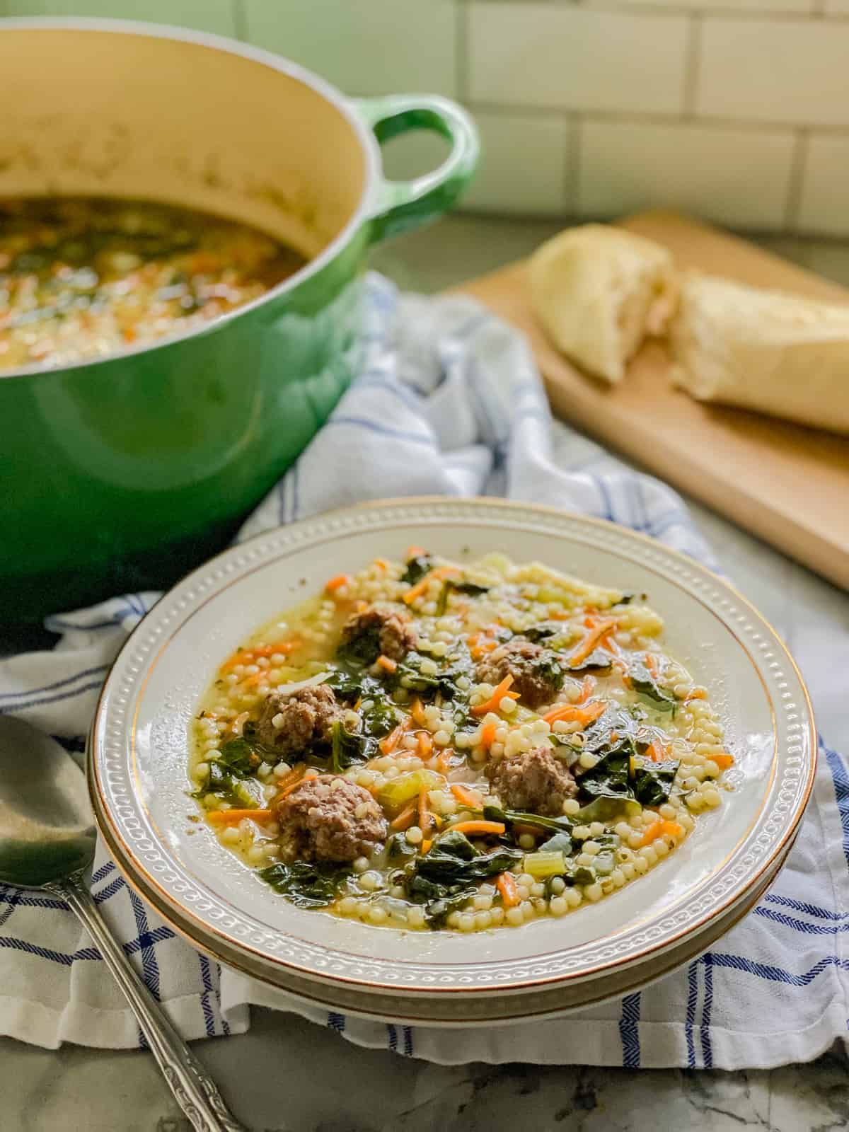 White bowl filled with meatball soup with green pot in background with a cutting board with a loaf of bread.