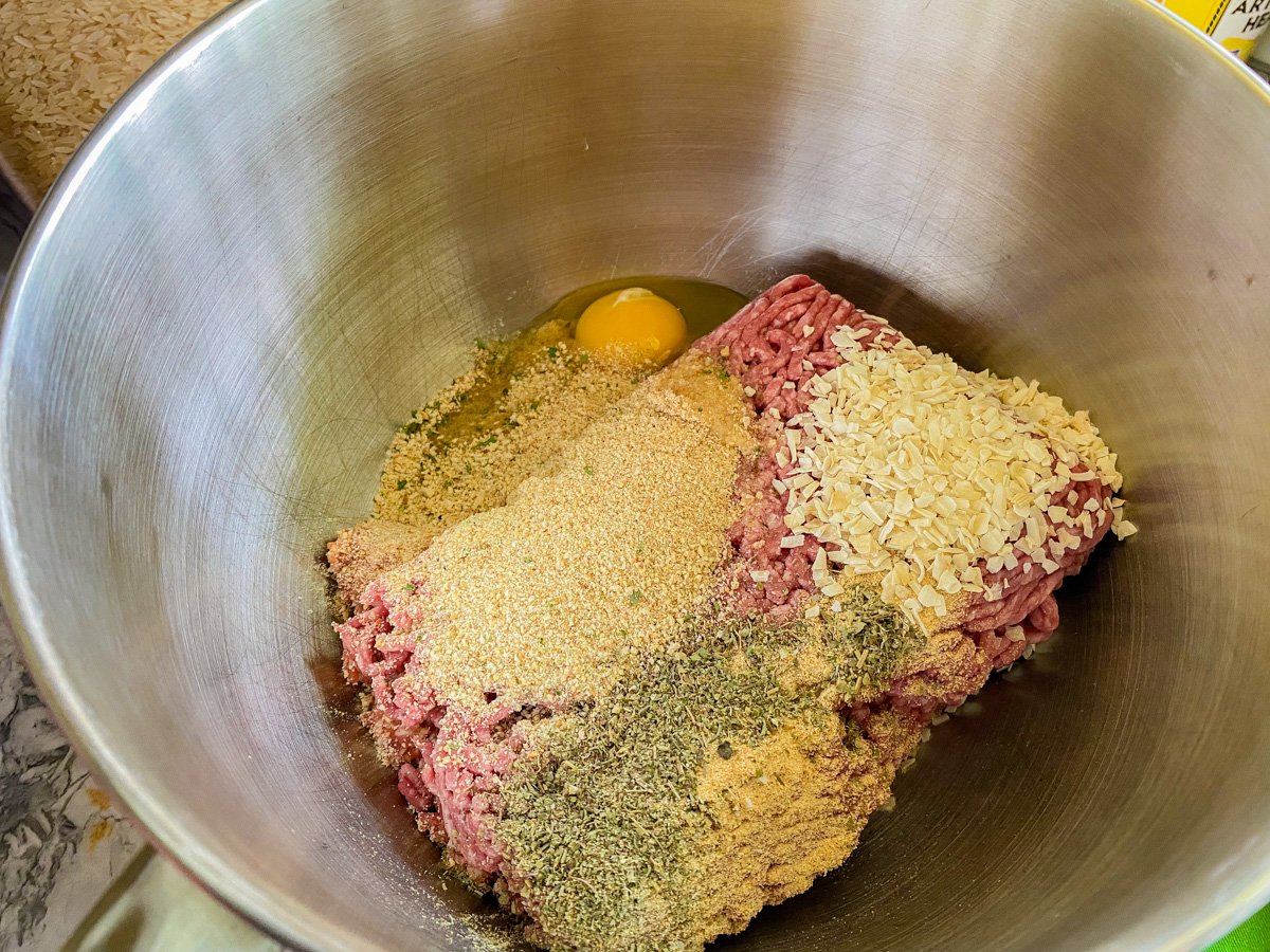 Ground beef and pork in a silver mixing bowl with spices, breadcrumbs, and egg in the bowl.
