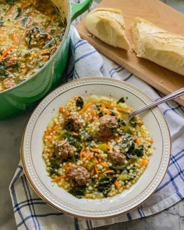 Bowl of meatball and pasta soup on a striped cloth with bread and a big pot of soup on the far left.