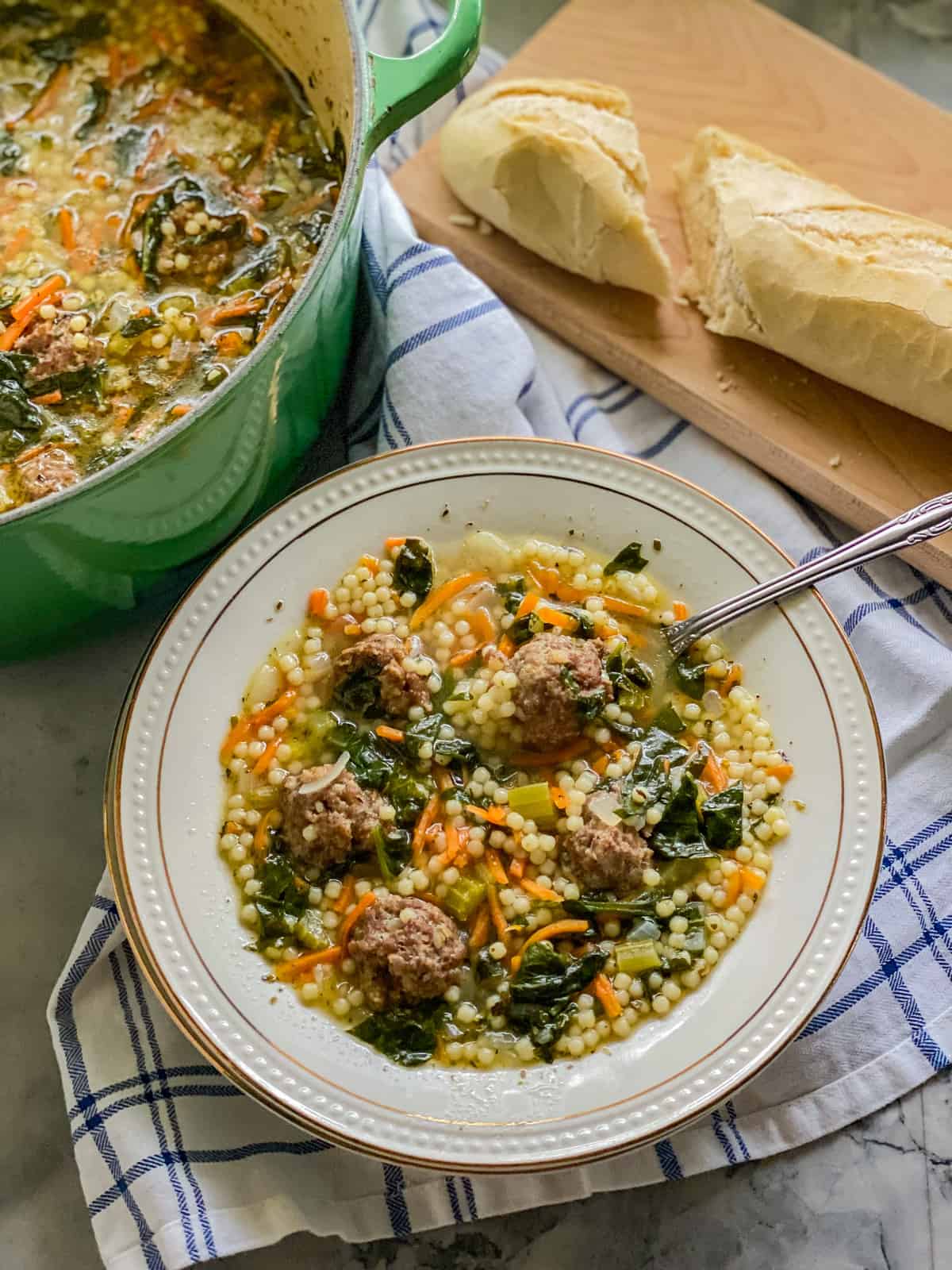 Bowl of meatball and pasta soup on a striped cloth with bread and a big pot of soup on the far left.